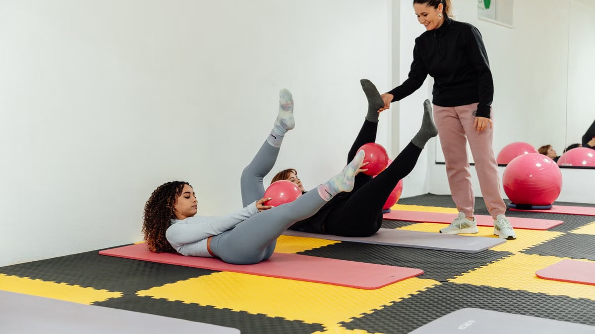 Women engaged in pilates class using pink balls and yoga mats indoors at a gym.